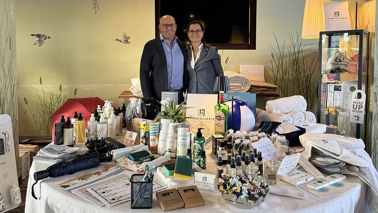Le Hotellerie: Qualità per Hotel a Lago di Garda: Un uomo e una donna sono in piedi dietro un tavolo rotondo su cui sono esposti vari articoli da hotel, tra cui prodotti da bagno, pantofole e prodotti di marca.