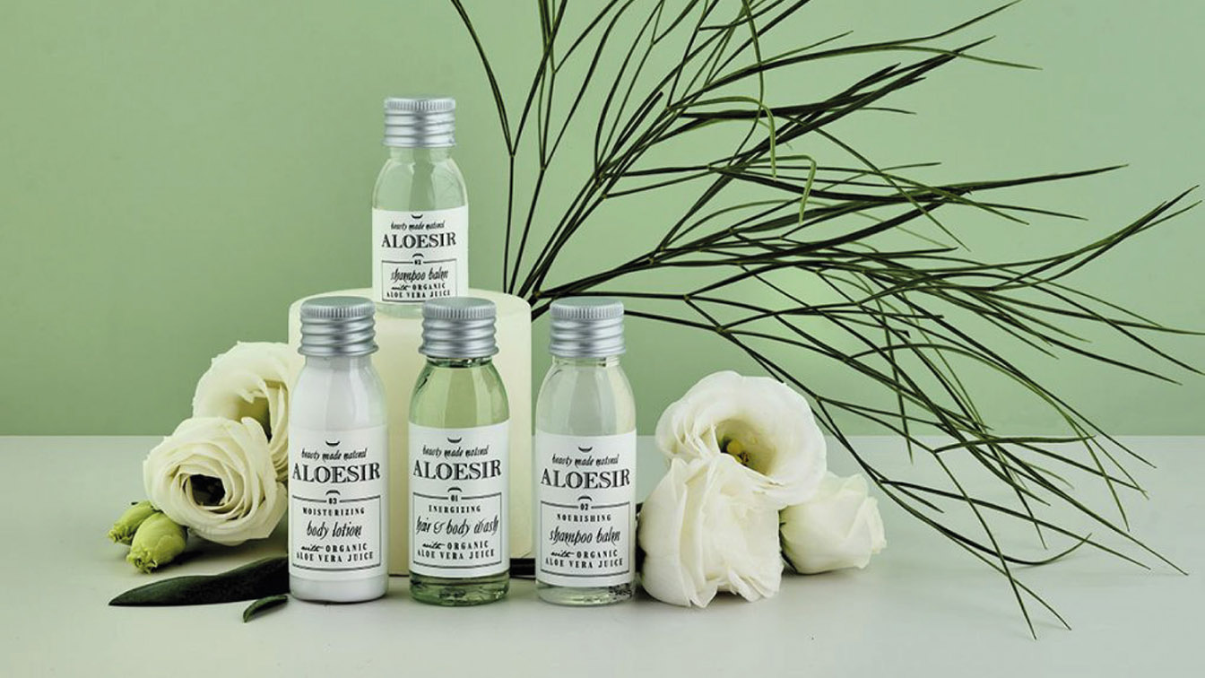 Coprimaterassi Usa e Getta Blu Service - Lago di Garda: Quattro flaconcini da viaggio Aloesir con le diciture lozione per il corpo, bagnoschiuma per capelli e corpo e balsamo shampoo, disposti insieme a fiori bianchi e foglie verdi.
