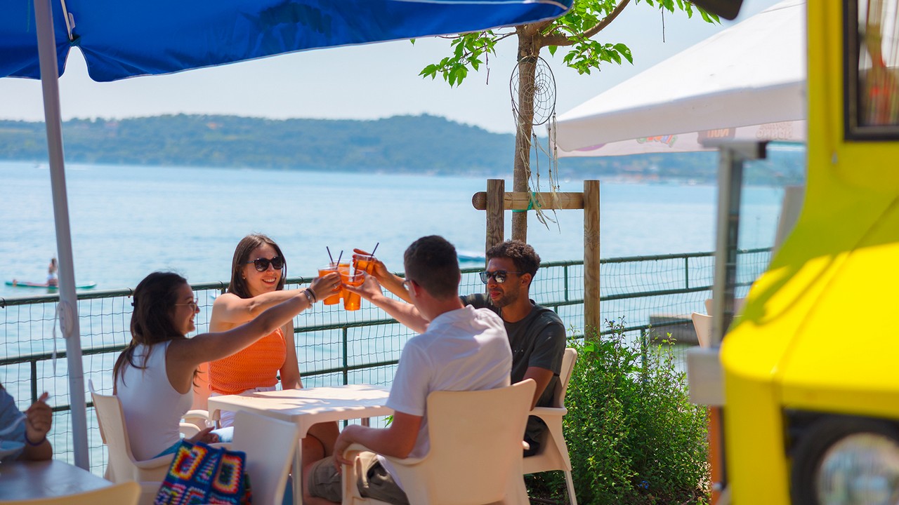 Offerta Speciale al Camping Riva Blu Lago di Garda: Quattro amici sono seduti a un tavolo all’aperto in riva al lago e brindano con delle bevande all’arancia.